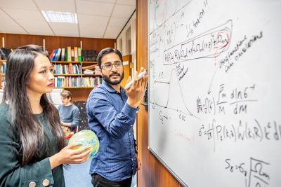 A physics lecturer writing on a whiteboard, presenting to a group of students