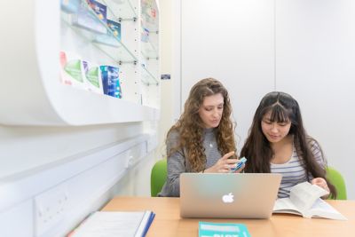 Two students sat in front of laptop one reading information on packet of tablets. Other student looking details up in a directory.
